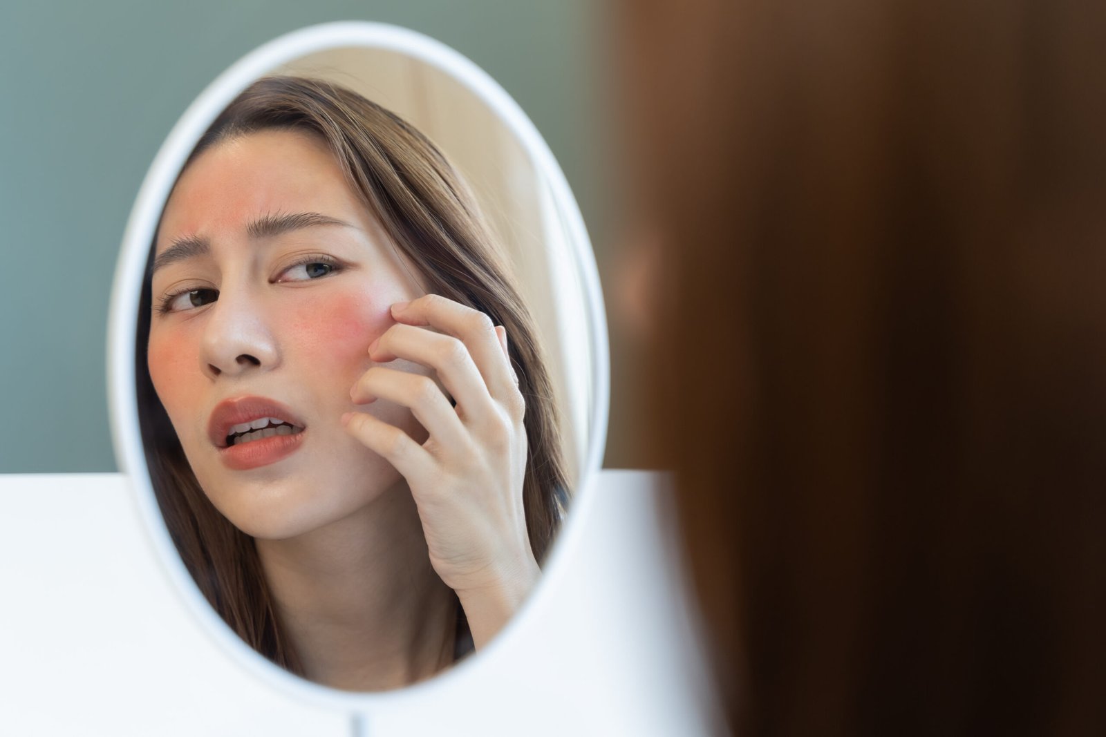 Asian young woman looking face skin in the mirror have a red rash on her face from cosmetic allergy