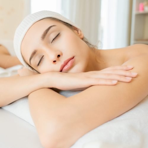 Close-up of a young woman relaxing at a spa, lying on a white towel with eyes closed, wearing a white headband, expressing peace and wellness during facial or body care treatment.