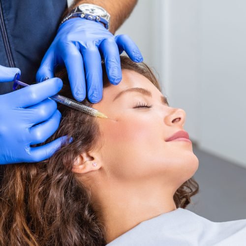 Attractive young woman is getting a rejuvenating facial injections. She is sitting calmly at clinic. The expert beautician is filling female wrinkles by hyaluronic acid.