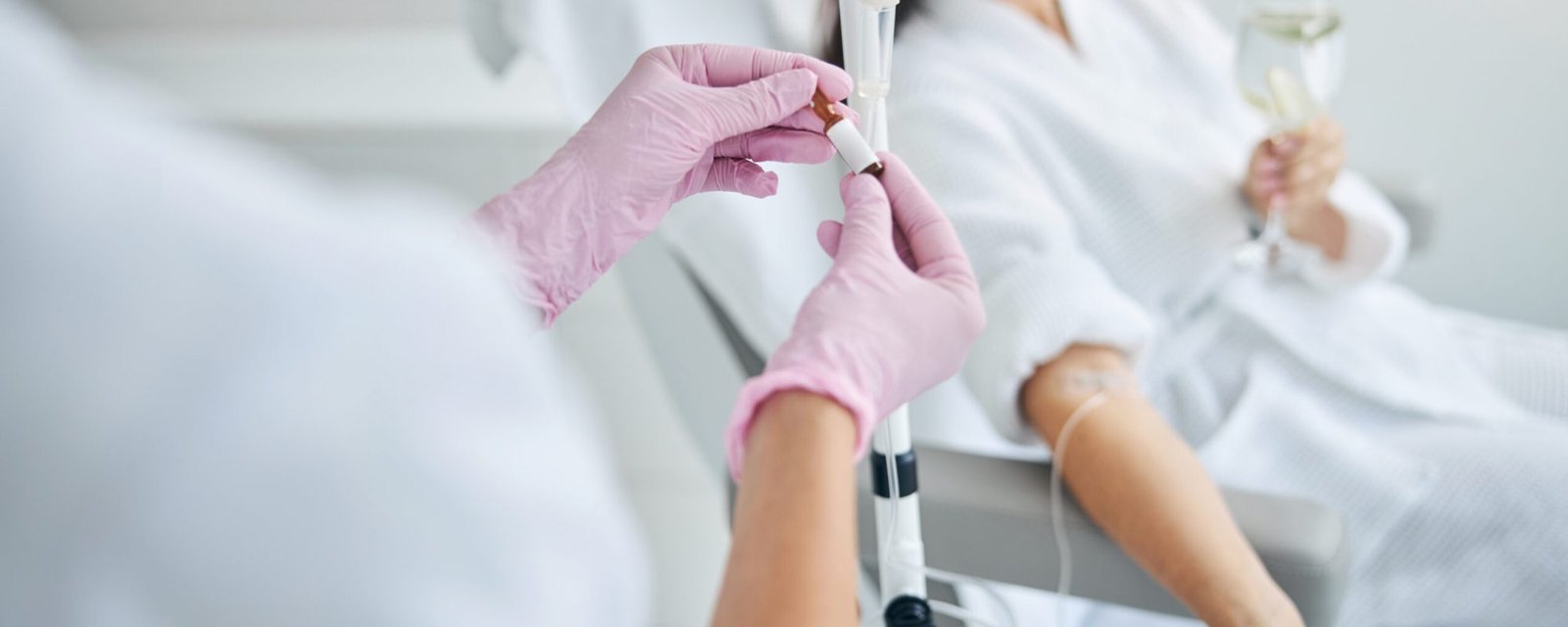 Nurse in latex gloves performing a medical procedure Dark-haired spa client with a glass of drink in her hand looking away during the intravenous therapy